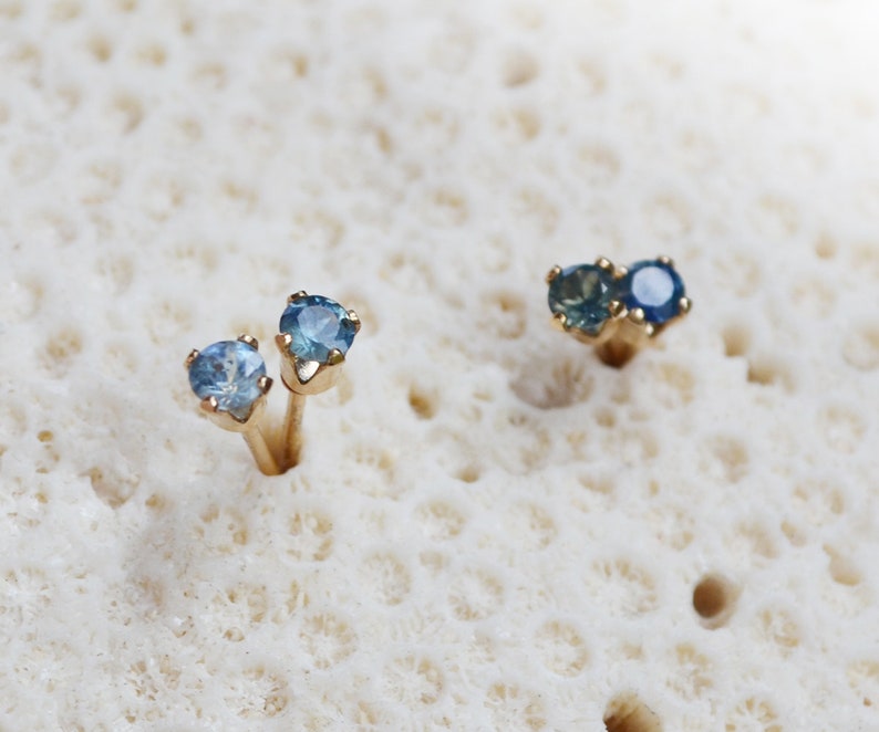 Puede incluir: Un par de pendientes de bot&oacute;n de oro con piedras preciosas azules. Los pendientes est&aacute;n colocados sobre un fondo blanco.