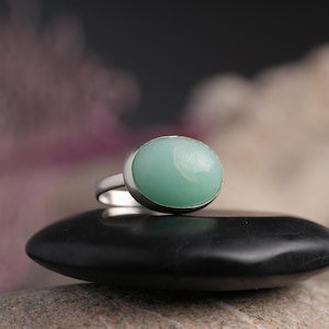 May include: A silver ring with a large, oval, light green gemstone. The ring is set on a black, smooth stone, with a blurred background of pink and white. The ring's band is thin and silver.