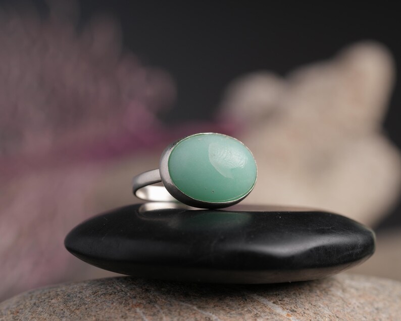 May include: A silver ring with a large, oval, light green gemstone. The ring is set on a black, smooth stone, with a blurred background of pink and white. The ring's band is thin and silver.
