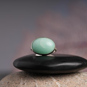 May include: A silver ring featuring a large, oval-shaped, pale green gemstone. The ring is set on a black, smooth stone, with a blurred background. The gemstone has a polished, smooth surface. The ring is a piece of jewelry.