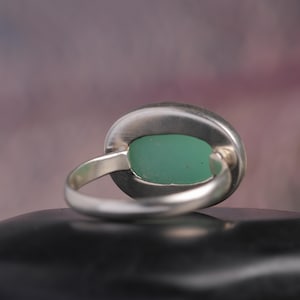May include: A silver ring with a large, oval-shaped, light green stone. The ring's band is a simple, polished silver, and the stone is set in a bezel setting. The ring is displayed on a black surface.