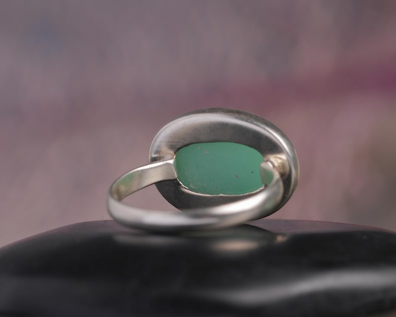May include: A silver ring with a large, oval-shaped, light green stone. The ring's band is a simple, polished silver, and the stone is set in a bezel setting. The ring is displayed on a black surface.