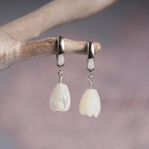 May include: A pair of dangle earrings with a floral design. The earrings feature a silver-colored hoop and a white, tulip-shaped pendant. The pendants are suspended by a silver wire. The earrings are displayed on a wooden branch against a soft, blurred background.