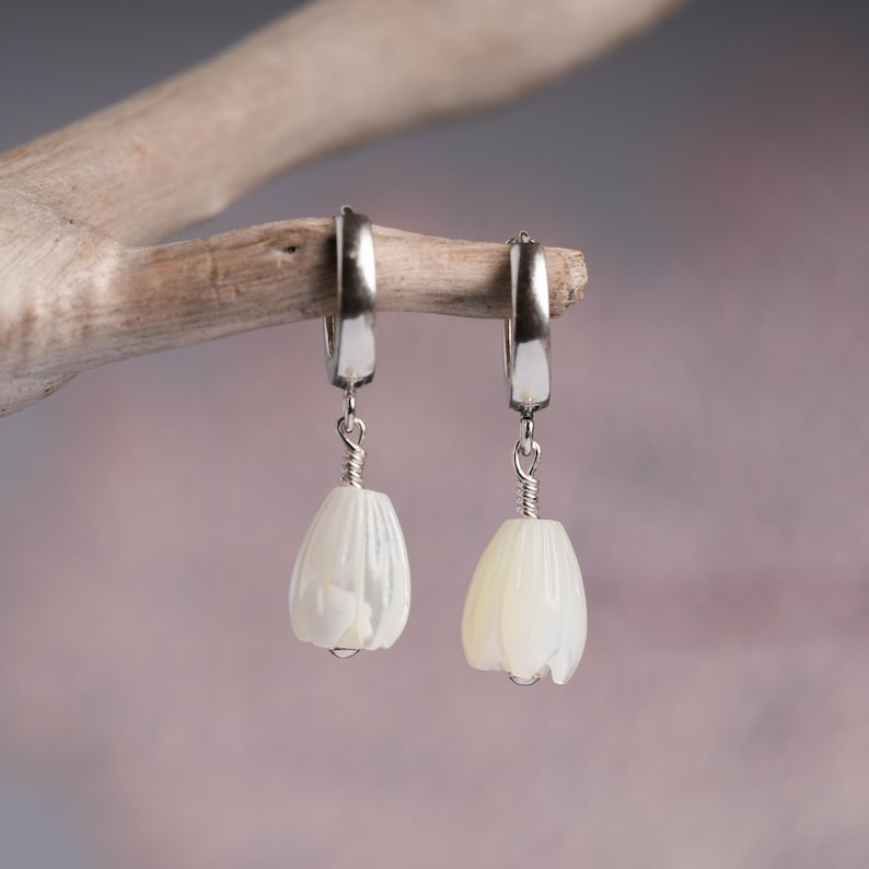 May include: A pair of dangle earrings with a floral design. The earrings feature a silver-colored hoop and a white, tulip-shaped pendant. The pendants are suspended by a silver wire. The earrings are displayed on a wooden branch against a soft, blurred background.