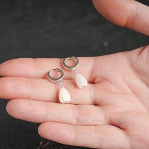 May include: A pair of silver hoop earrings with white, teardrop-shaped pendants. The earrings are displayed on a person's open palm against a dark background. The pendants have a textured surface.