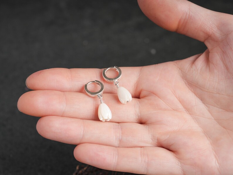 May include: A pair of silver hoop earrings with white, teardrop-shaped pendants. The earrings are displayed on a person's open palm against a dark background. The pendants have a textured surface.