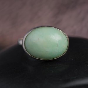 May include: A silver ring featuring a large, oval-shaped, pale green gemstone. The gemstone is set in a simple, polished silver band. The ring is displayed on a dark surface, with a blurred background.
