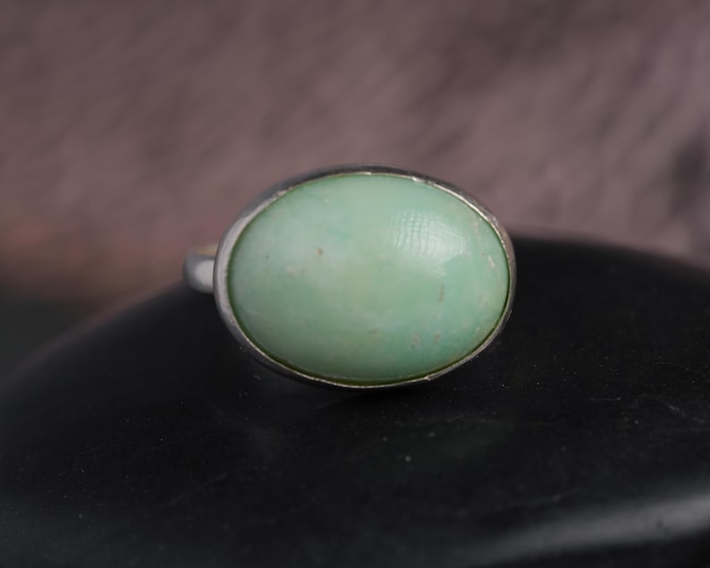 May include: A silver ring featuring a large, oval-shaped, pale green gemstone. The gemstone is set in a simple, polished silver band. The ring is displayed on a dark surface, with a blurred background.