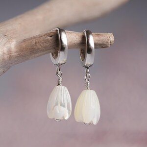 May include: A pair of silver hoop earrings with white, flower-shaped pendants. The pendants are suspended by a silver wire and have a pearlescent finish. The earrings are displayed on a piece of driftwood against a soft, blurred background.