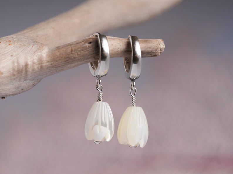 May include: A pair of silver hoop earrings with white, flower-shaped pendants. The pendants are suspended by a silver wire and have a pearlescent finish. The earrings are displayed on a piece of driftwood against a soft, blurred background.