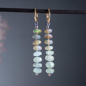 Peut inclure: Boucles d'oreilles créoles dorées avec des rangées de pierres précieuses ovales et facettées dans des tons de vert, d'opale et de bleu. Les boucles sont suspendues à une tige sombre sur fond gris.