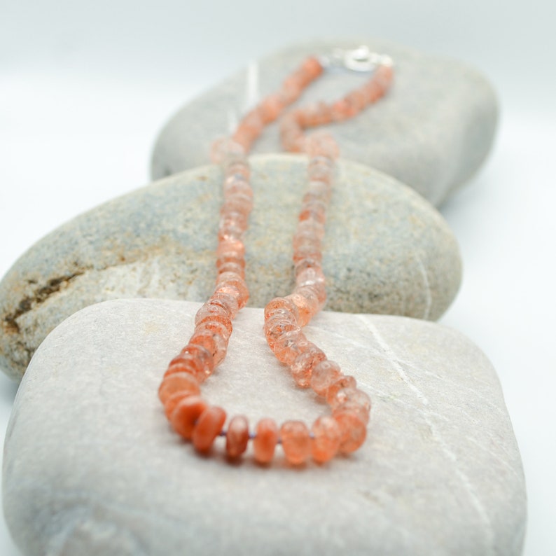 May include: A necklace made of small, faceted, peach-colored sunstone beads. The necklace is displayed on a bed of smooth, gray stones.