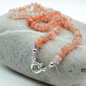 May include: A necklace made of small, light orange sunstone beads with a silver clasp. The necklace is lying on a gray stone.