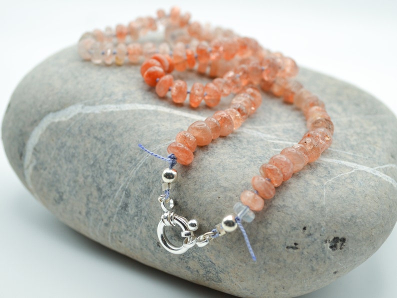 May include: A necklace made of small, light orange sunstone beads with a silver clasp. The necklace is lying on a gray stone.
