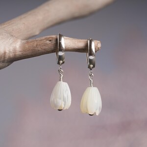 May include: A pair of silver hoop earrings with white, teardrop-shaped pendants. The pendants are carved with a petal design and have a small, dark bead at the bottom. The earrings are displayed on a light brown branch against a soft, blurred background.