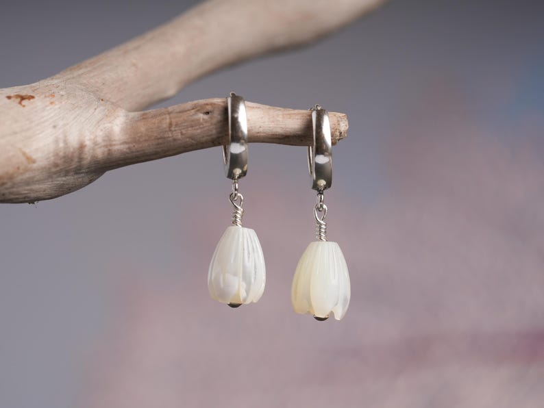 May include: A pair of silver hoop earrings with white, teardrop-shaped pendants. The pendants are carved with a petal design and have a small, dark bead at the bottom. The earrings are displayed on a light brown branch against a soft, blurred background.