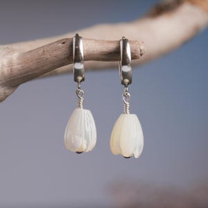 May include: A pair of white, flower-shaped earrings with silver-colored hoops and wire detailing. The earrings are hanging from a wooden branch against a blurred blue and gray background. The earrings have a pearlescent sheen.