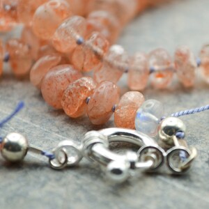 May include: A close-up of a sunstone necklace with a silver clasp. The necklace is made of orange and red sunstone beads and is strung on a blue cord.