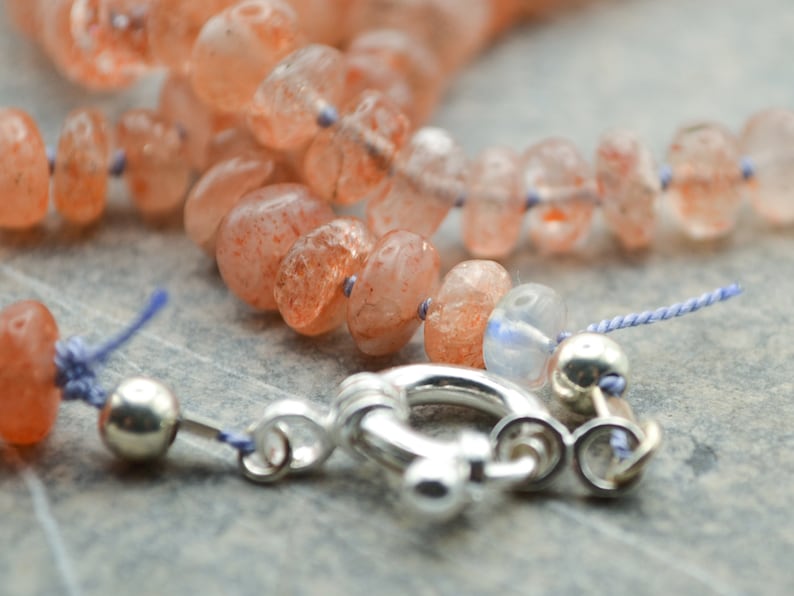 May include: A close-up of a sunstone necklace with a silver clasp. The necklace is made of orange and red sunstone beads and is strung on a blue cord.