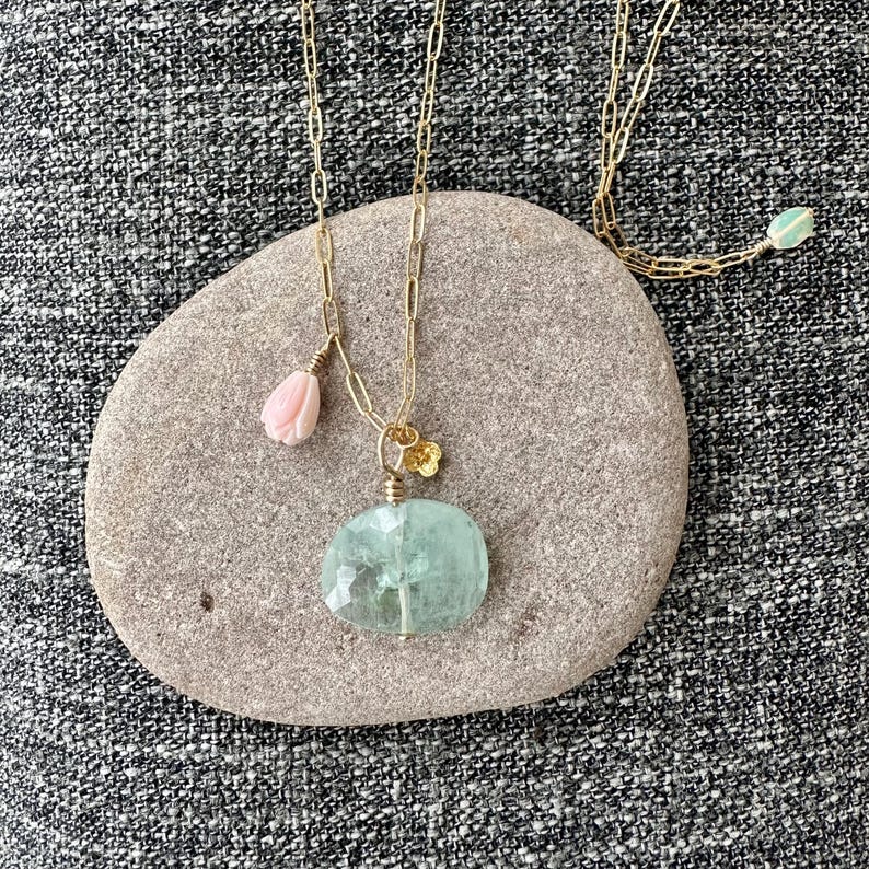 May include: A gold chain necklace with a large, light blue aquamarine pendant, a small pink flower charm, and a small gold flower charm.