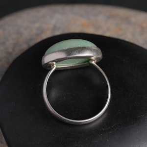May include: A silver ring with a light green, oval-shaped stone. The stone is set in a silver bezel and the band is a simple, polished silver. The ring is displayed on a dark, smooth surface.