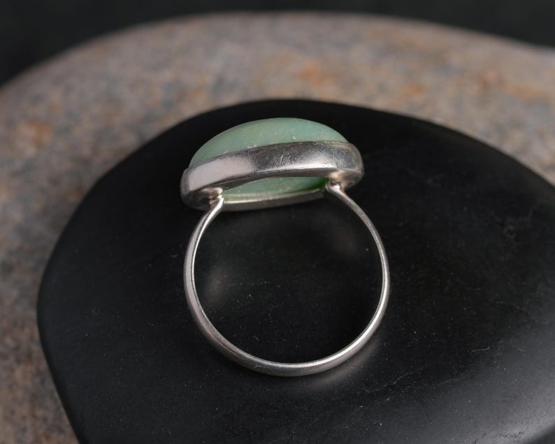 May include: A silver ring with a light green, oval-shaped stone. The stone is set in a silver bezel and the band is a simple, polished silver. The ring is displayed on a dark, smooth surface.