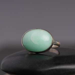 May include: A silver ring featuring a large, oval-shaped, pale green gemstone. The stone is set in a simple, polished silver band. The ring is displayed on a black surface against a gray background, highlighting the ring's design.