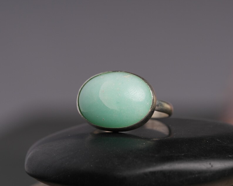 May include: A silver ring featuring a large, oval-shaped, pale green gemstone. The stone is set in a simple, polished silver band. The ring is displayed on a black surface against a gray background, highlighting the ring's design.