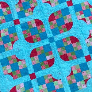 May include: A colorful quilt with a repeating pattern of squares and rounded shapes. The quilt features squares of red, blue, green, and pink fabric on a light blue background. The quilt has a stitched dragonfly pattern.