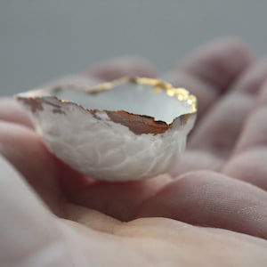 May include: A small, white ceramic bowl with a gold rim. The bowl has a textured surface and is held in a hand.