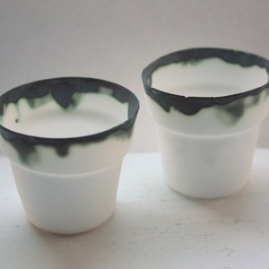 Puede incluir: Dos tazas de cerámica blanca con un borde verde oscuro y negro. Las tazas están vacías y están sobre una superficie blanca.