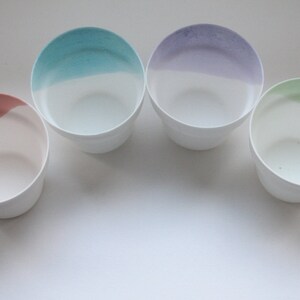 May include: Four ceramic bowls with a white interior and a coloured rim. The bowls are arranged in a semi-circle on a white surface. The colours of the rims are pink, blue, purple, and green.