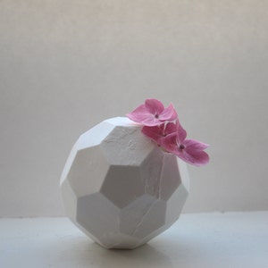 Puede incluir: Un jarrón geométrico blanco con un arreglo de flores rosas. El jarrón está hecho de cerámica y tiene una superficie texturizada. El arreglo floral está hecho de hortensias rosas.