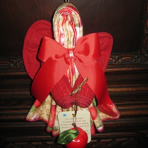 May include: A red and white kitchen towel angel ornament with a red bow and a green apple with a red stem. The ornament has a tag that reads "I'm a kitchen angel to hang up on your wall. I'll help you with your baking. What you see that makes don't..."