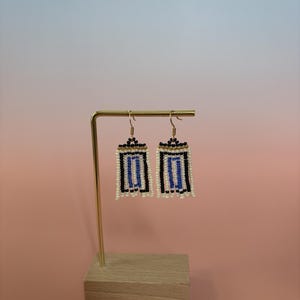May include: A pair of rectangular beaded earrings featuring a geometric pattern of white, black, and blue seed beads. The earrings are suspended from gold-toned metal hooks and have a fringe of small white beads along the bottom edge.