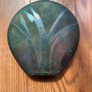 May include: A round, flat decorative object with a textured, dark green and bronze patina finish. The surface features an embossed, stylized plant design with long, slender leaves. The name William is inscribed in gold script at the bottom edge.