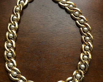 May include: A gold-toned necklace with a chain-link design, interspersed with small, round, white faux pearls. The necklace is unclasped, showing the clasp mechanism. The jewelry is displayed on a dark brown wooden surface.