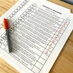 May include: A printed checklist titled 'Minimize & Organise' with 365 daily prompts, handwritten numbers, and an orange marker. The prompts encourage decluttering and organisation throughout the home.