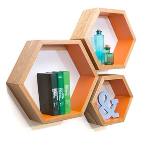 May include: Three hexagonal shelves made of ash wood with a natural finish and a bright orange paint. The shelves are stacked on top of each other and are mounted on a white wall. The top shelf has two glass bottles, the middle shelf has three books, and the bottom shelf has a blue ampersand.