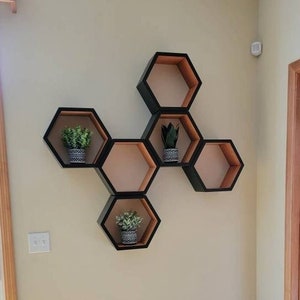 May include: A set of six hexagonal shelves in black and wood tones, arranged in a honeycomb pattern on a white wall. The shelves are decorated with small potted plants.