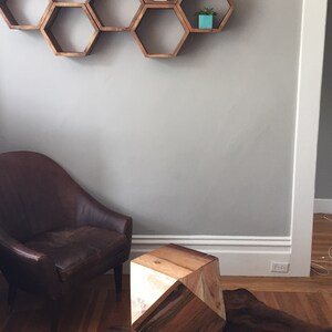 May include: A wooden hexagonal shelf unit with six compartments, mounted on a light grey wall. The shelf is decorated with small potted plants. A brown leather armchair sits in front of the shelf, and a wooden geometric stool sits on a cowhide rug.