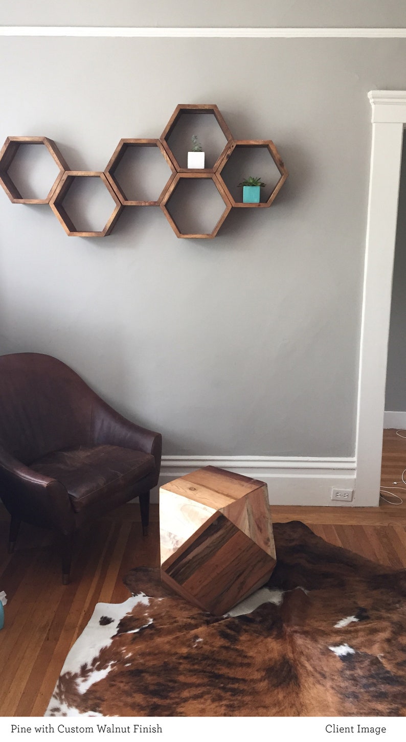 May include: A wooden hexagonal shelf unit with six compartments, mounted on a light grey wall. The shelf is decorated with small potted plants. A brown leather armchair sits in front of the shelf, and a wooden geometric stool sits on a cowhide rug.