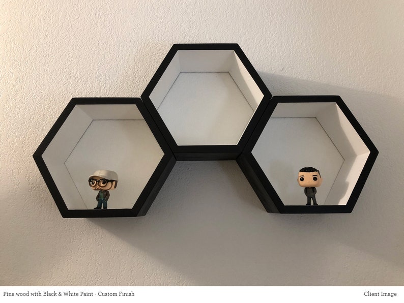 May include: Three hexagonal shelves made of pine wood, painted black and white, are mounted on a white wall. Two small figurines are displayed on the shelves.