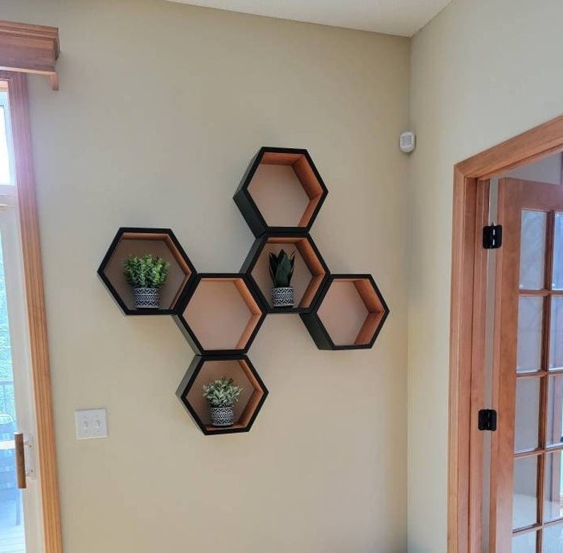 May include: A set of six hexagonal shelves in black and wood tones, arranged in a honeycomb pattern on a white wall. The shelves are decorated with small potted plants.