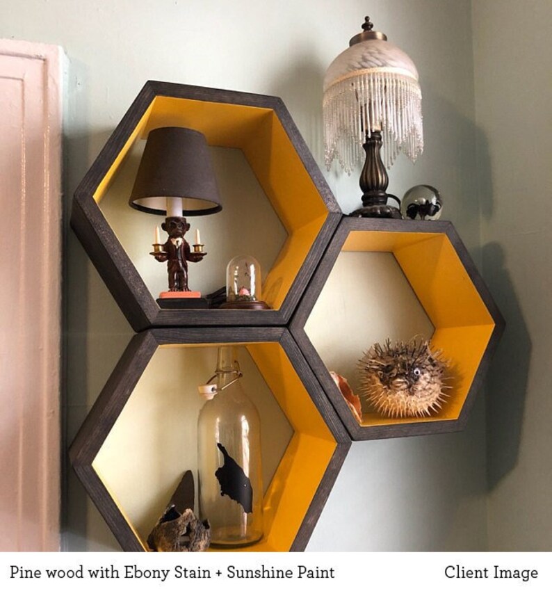 May include: A set of hexagonal shelves made from pine wood, stained in ebony and painted in sunshine yellow. The shelves are arranged in a honeycomb pattern and feature various decorative items, including a lamp with a black shade, a glass bottle, a small figurine, and a pufferfish.