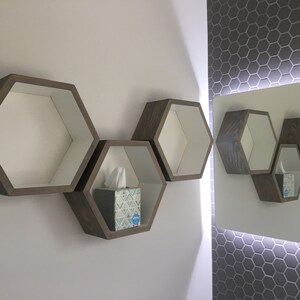 May include: Three wooden hexagonal shelves mounted on a white wall. The shelves are empty except for the middle shelf, which has a box of tissues on it. The shelves are reflected in a mirror on the wall to the right.