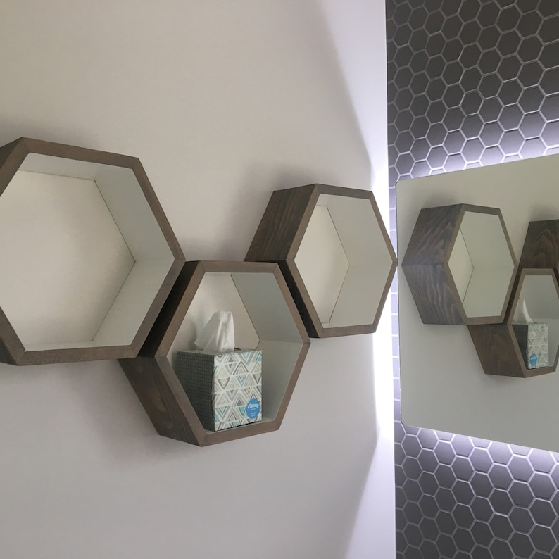 May include: Three wooden hexagonal shelves mounted on a white wall. The shelves are empty except for the middle shelf, which has a box of tissues on it. The shelves are reflected in a mirror on the wall to the right.