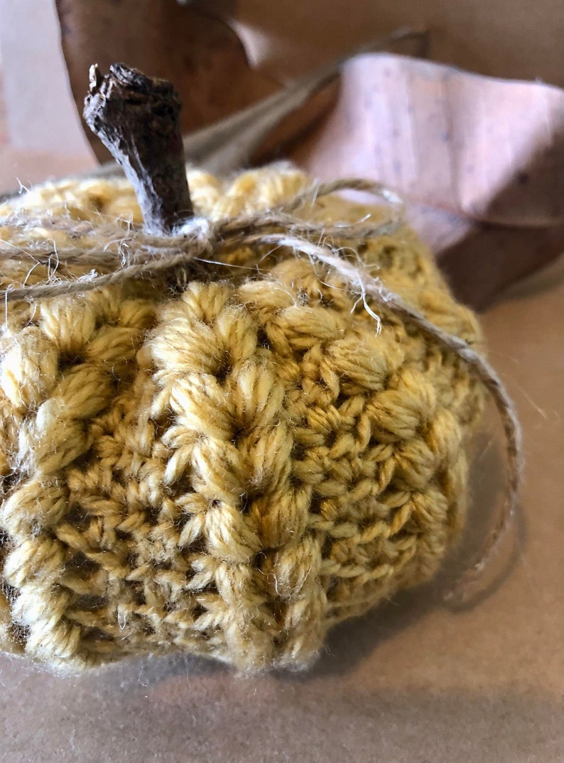 May include: A crocheted pumpkin in a yellow color with a brown twig stem and twine tied around the top.
