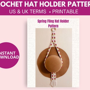 May include: A crochet hat holder pattern with instructions for a spring fling hat holder. The pattern includes US and UK terms and is printable. The image shows a brown hat hanging on the holder, which is made with white yarn and pink flowers.