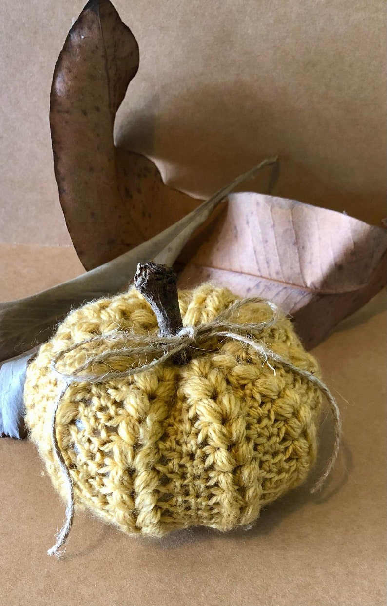 May include: A crocheted pumpkin in a yellow color with a twig stem and twine tied around the stem. The pumpkin is sitting on a brown surface.